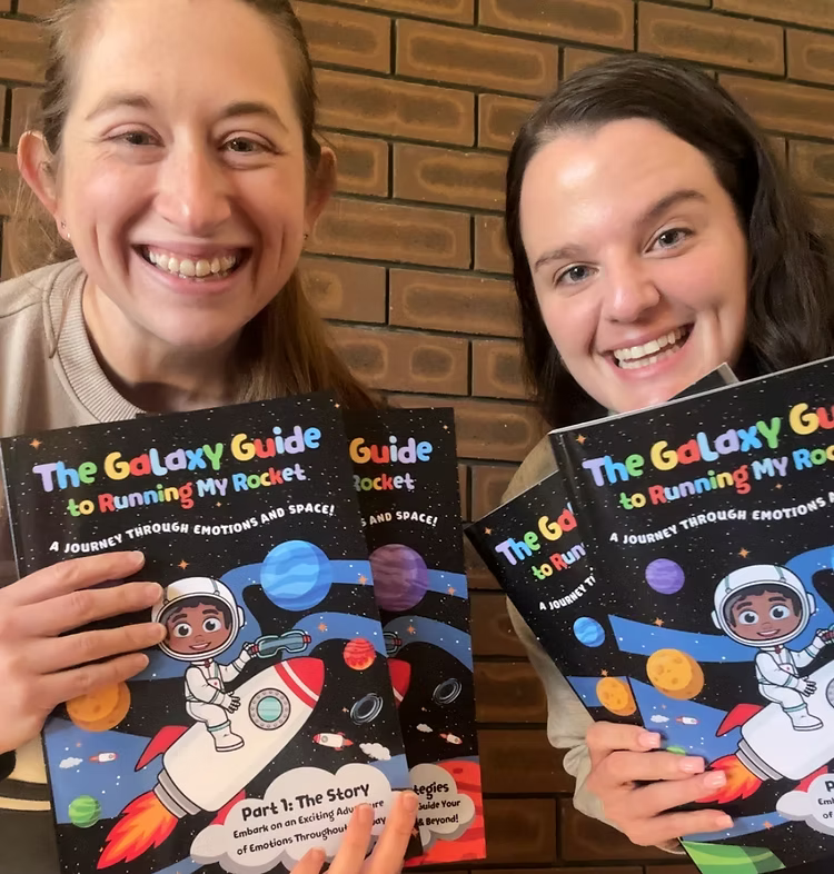 Two therapists holding books titled 'The Galaxy Guide to Running My Rocket' against a brick wall.
